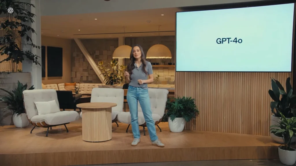 Mira Murati présentant devant un écran affichant 'GPT-4o' dans une salle moderne avec des fauteuils et des plantes.